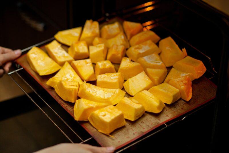 a partial image of a couple of hands putting a baking tray in the oven full of cubed pumpkin ready for baking