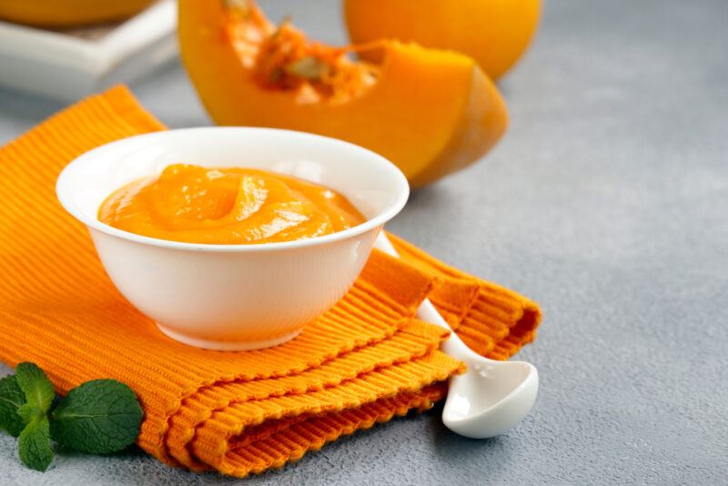 on a grey concrete surface is a white bowl full of pumpkin puree resting on top of an orange table napkin with pumpkin slices at the back