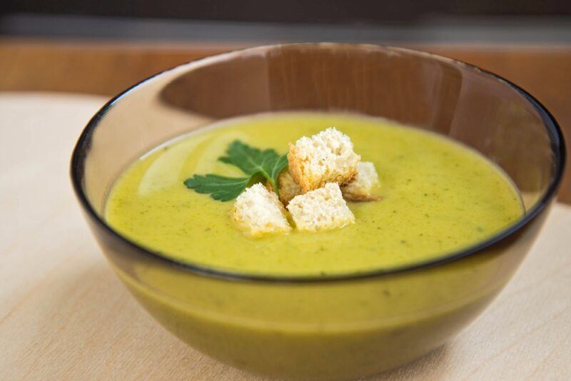 a closeup image of a glass bowl with pea puree soup with croutons and herbs on top