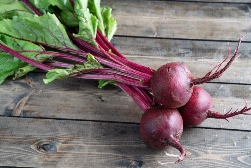 on a wooden surface are three purple beets with leaves still attached