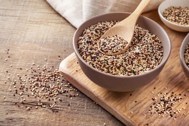 on a wooden surface is a wooden chopping board with brown bowl full raw quinoa, at the back is a beige table napkin, and around are loose quinoa grains