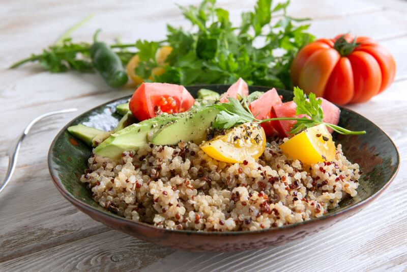 on a marble surface is black glass dish with quinoa salad with cut tomatoes and herbs on top, at the back is a bunch of cilantro  with a whole red tomato