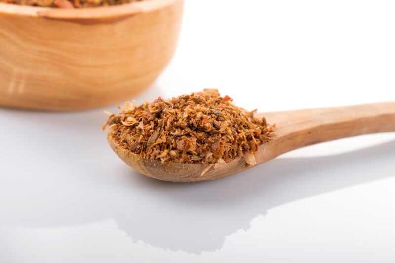 on a white surface is a wooden spoon full of ras el hanout, a partial image of a wooden bowl with ras el hanout can be seen at the back