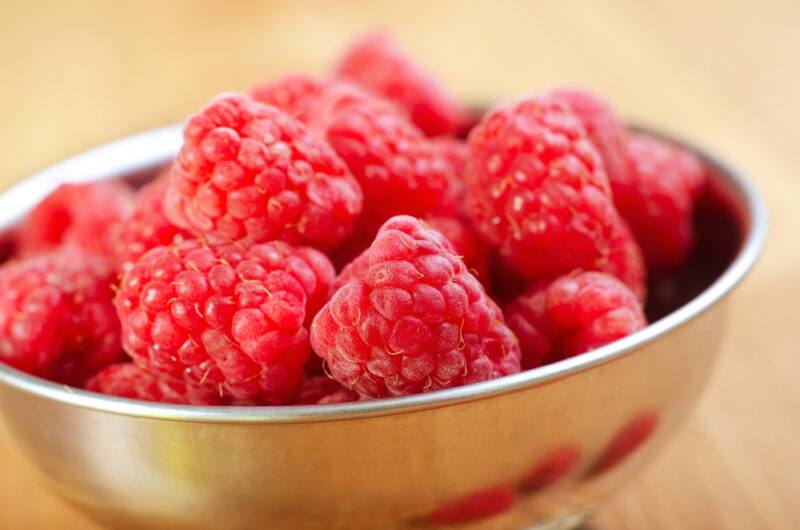 on a wooden surface is a closeup shot of a metal bowl full of raspberries