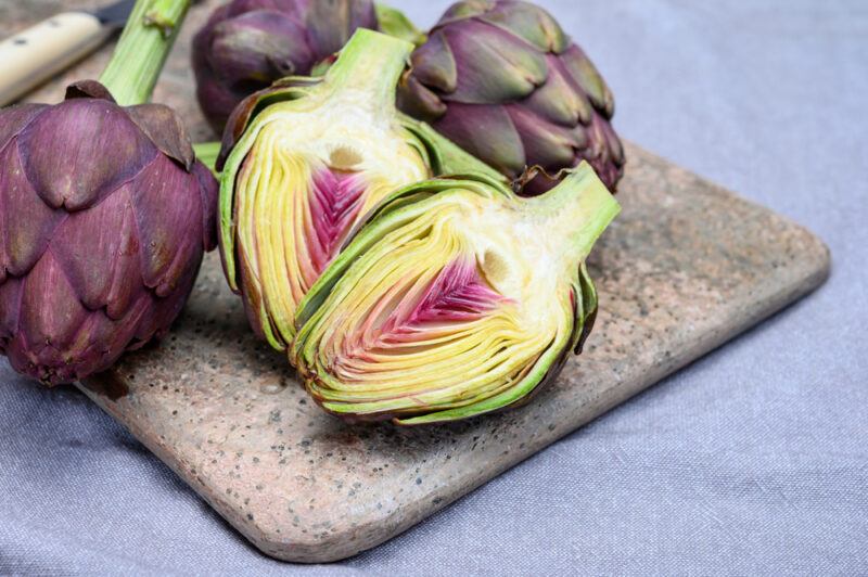 bluish table cloth with a wooden chopping board with halve and whole raw artichoke