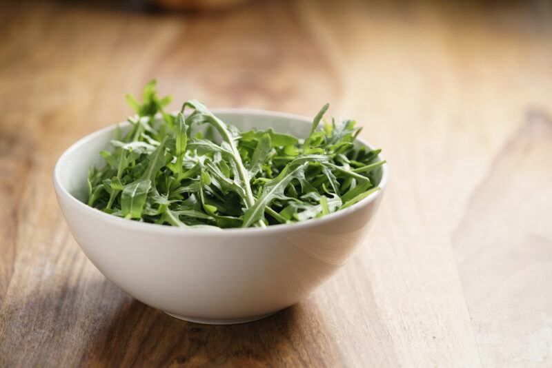 on a wooden surface is a white bowl full of raw arugula