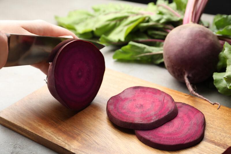 hands slicing raw beets into thin slices on a a wooden chopping board with one whole beat and green leaves at the back