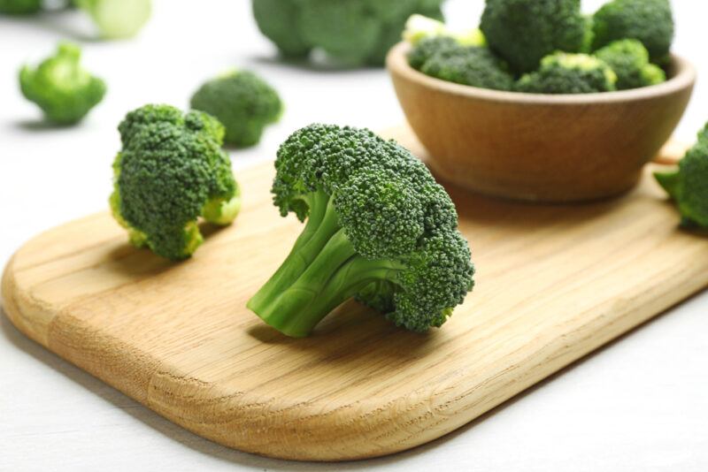 a closeup image of a wooden chopping board with broccoli florets with a small bowl full of broccoli florets, around the chopping board are loose florets as well