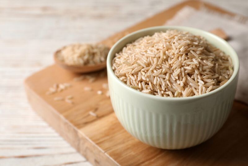 on a wooden surface is a wooden chopping board with a bowl of raw brown rice with a wooden spoon beside it full of raw brown rice as well