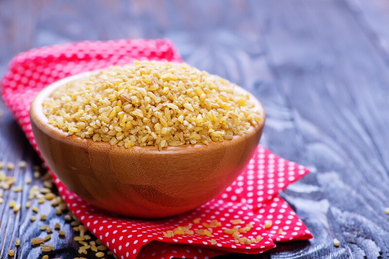 rustic wooden surface with a brown wooden bowl full of raw bulgar resting on top of a pink table napkin with white dots