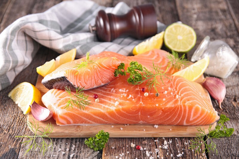 on an aged wooden surface is a closeup image of raw salmon fish on a wooden board with herbs and spices and sliced lemon, with wooden pepper grinder and kitchen towel