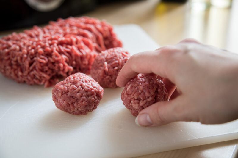 raw ground beef on a white plastic chopping board with a couple of meatballs, a hand holding on to one of the meatballs can be seen