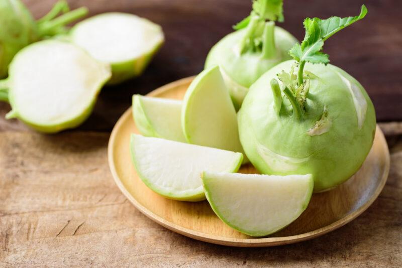 on a wooden surface is a light brown wooden plate with cut and whole kohlrabi with halve kohlrabi at the back