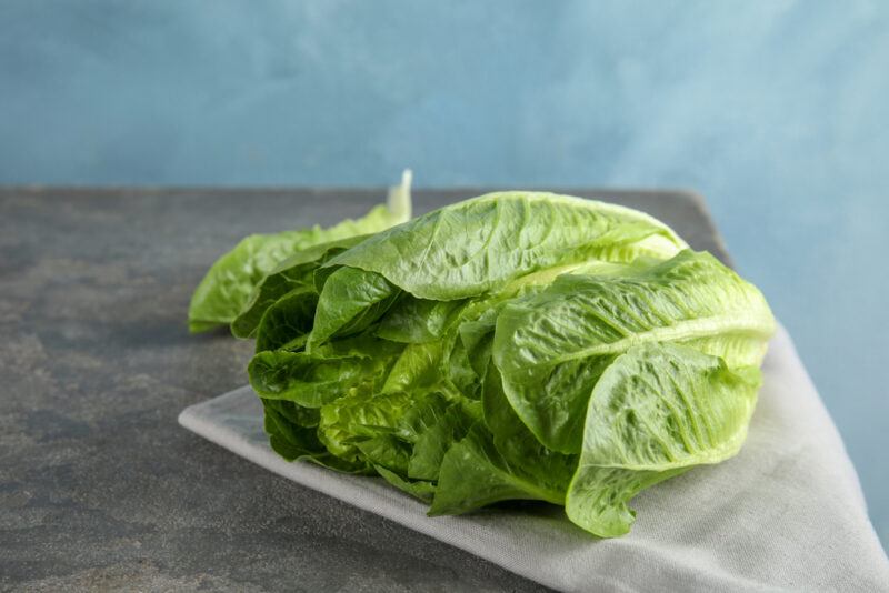 on a wooden table is tan-colored table napkin near the edge with a head of lettuce resting on it