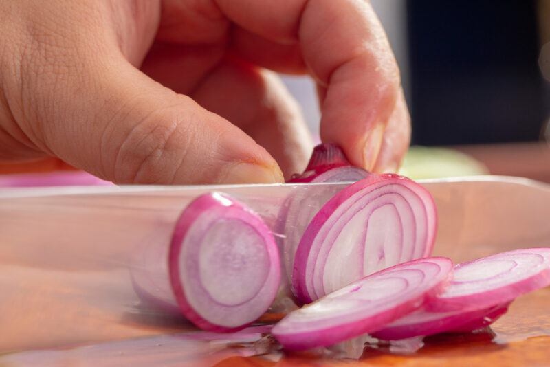 hands slicing red onion on a wooden chopping board