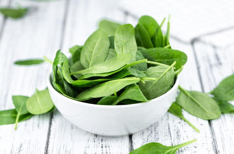 on a rustic white wooden surface is a white bowl full of raw spinach, with loose spinach around it