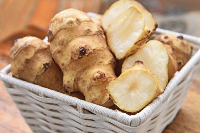 on a wooden surface is a closeup image of a tiny white basket full of raw whole and sliced sunchokes