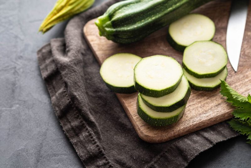 on a dark concrete surface is a dark table napkin underneath a wooden chopping board with sliced and whole zucchinis