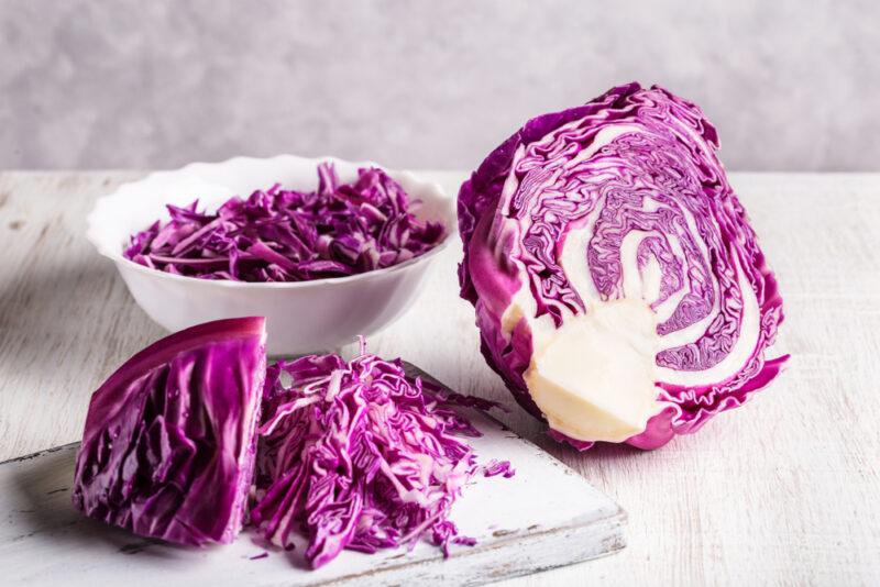 on a white wooden surface are halved red cabbage head, a white bowl of shredded red cabbage, and a wooden chopping board with shredded red cabbage on it