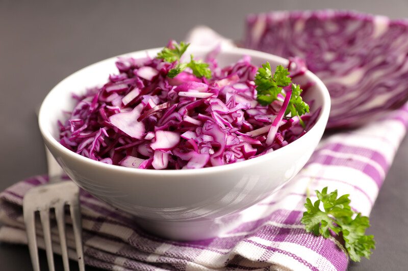 a white bowl full of shredded red cabbage with a sprig of herb, underneath it is a white and magenta table napkin with a sprig of herb and silver fork on its side and a slice of red cabbage at the back