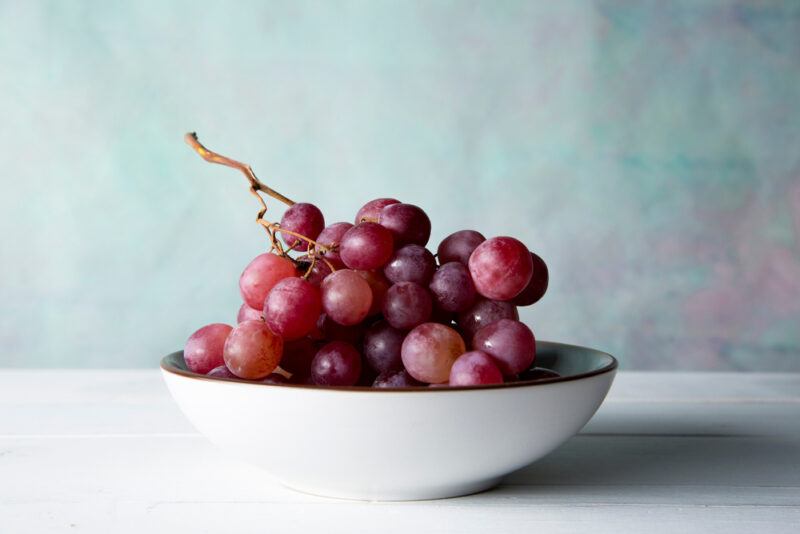 on a white surface and rustic looking bluish background is a white bowl full of red grapes 