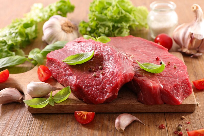 closeup image of a couple of red meat chunks resting on a wooden chopping board with herbs and spices on and around it