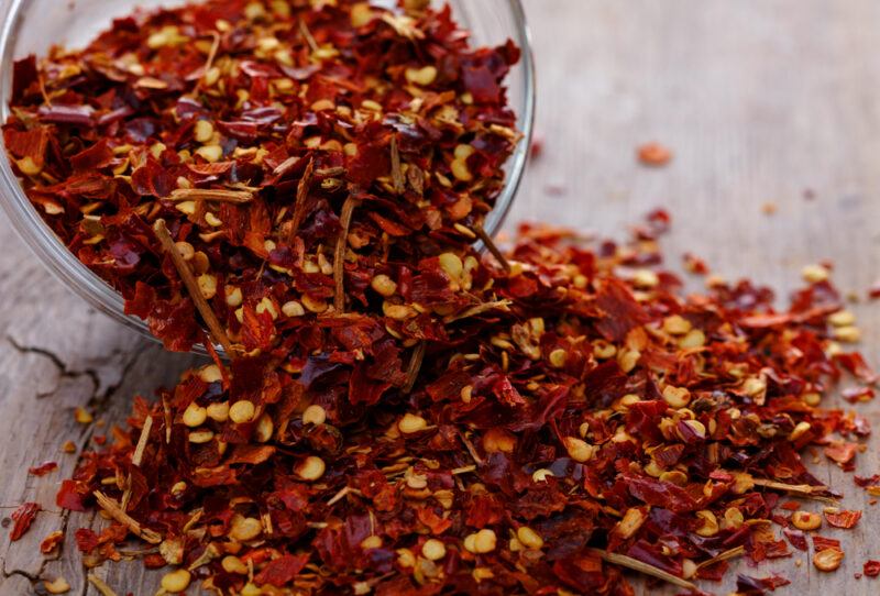 on a wooden surface is a closeup image of a mound of crushed red pepper flakes poured out from a jar beside it