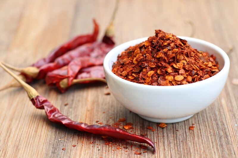 on a wooden surface is a white bowl full of red pepper flakes with loose pepper flakes and dried cayenne pepper around it