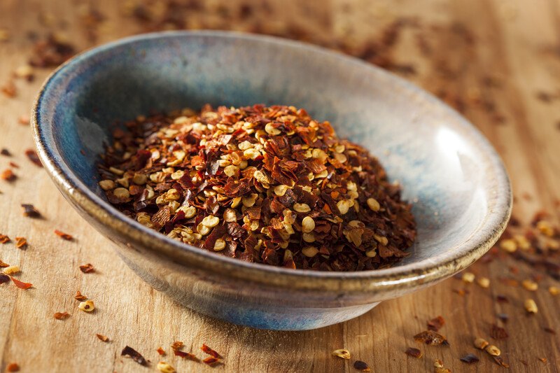 closeup image of a shallow grey-bluish bowl with red pepper flakes on a wooden surface with loose red pepper flakes around it