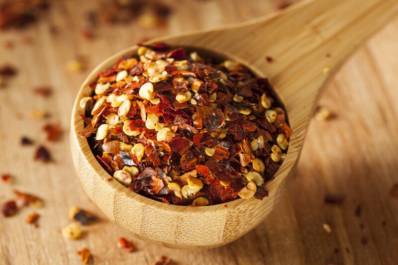 closeup image of a wooden scoop full of red pepper flakes on a wooden surface with loose red pepper flakes around it