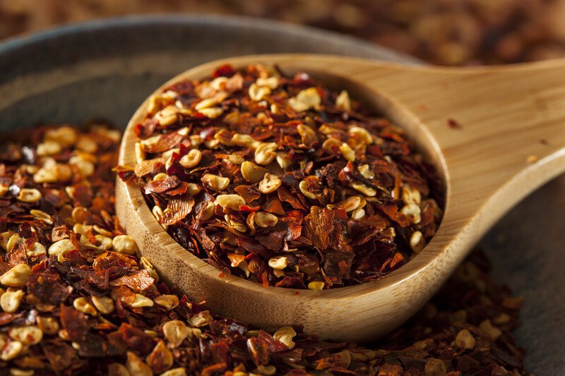 closeup image of a wooden scoop full of red pepper flakes resting on top of a mound of red pepper flakes