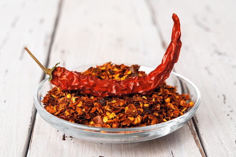 on a rustic wooden surface is a transparent dish full of red pepper flakes, with a dried chili pepper on top