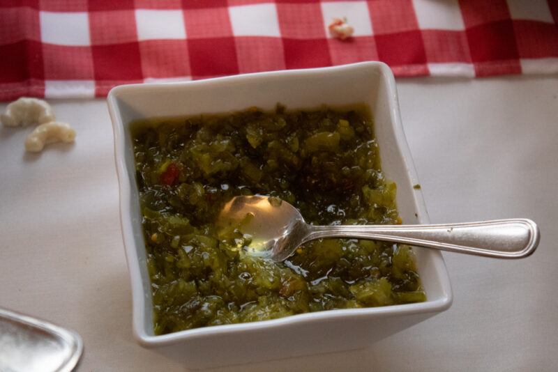 on a white surface is a white square dish with relish and a silver spoon, behind it is red and white table napkin