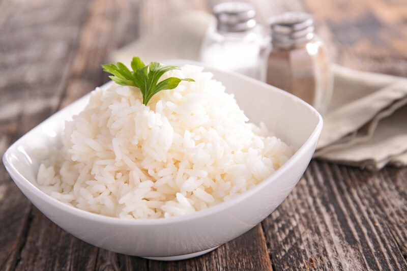 a white dish full of cooked white rice with a piece of parsley on top, at the back is a salt and pepper shaker on top of a tan-colored table napkin