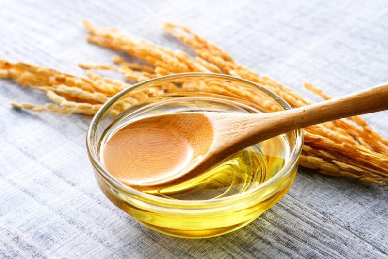 on a wooden surface is a small clear glass bowl full of rice bran oil with a wooden spoon in it, at the back is a rice stalk