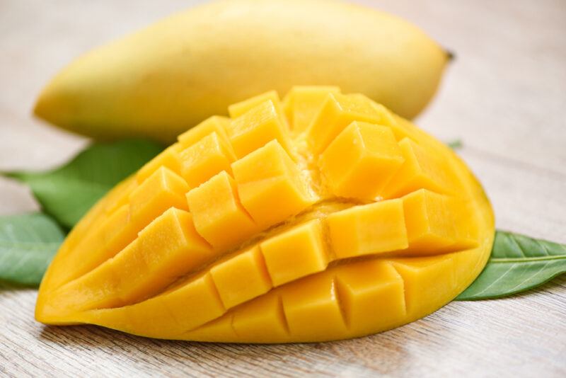 on a wooden surface is a closeup image of a sliced ripe mango resting on top of a couple of mango leaves, at the back is a whole ripe mango