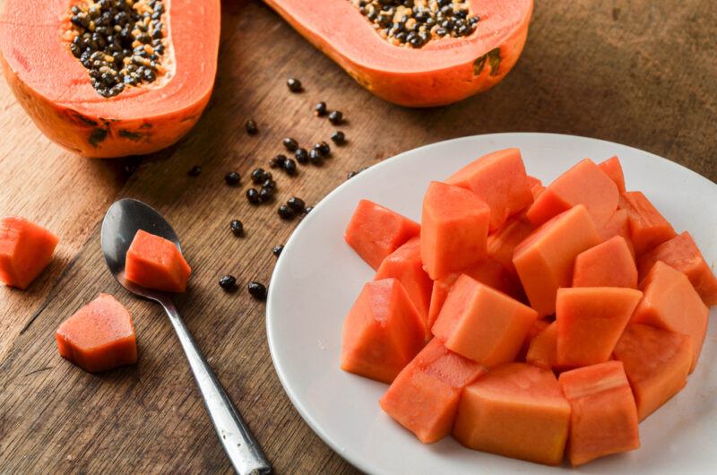 on a wooden surface is a white dish full of cubed yellow papaya, at the back is a halved ripe papaya with black seeds showing, around them are loose papaya seeds and cube ripe papaya