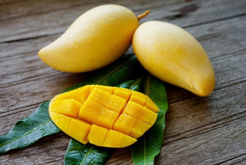 on a wooden surface are two whole ripe mangoes, and a sliced one resting on top of three mango leaves