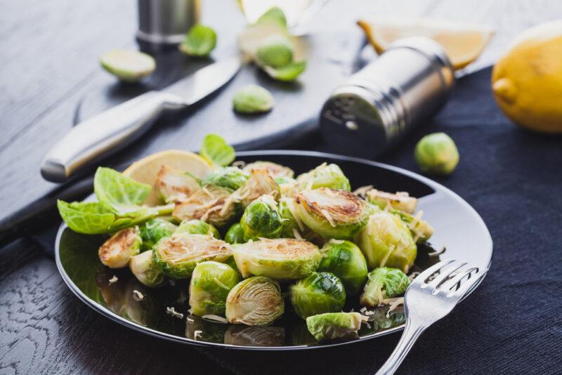 on a concrete surface is a black dish of roasted Brussels sprouts with silver fork and knife beside, around it are loose Brussels sprouts, glass salt shaker, and wooden pepper grinder