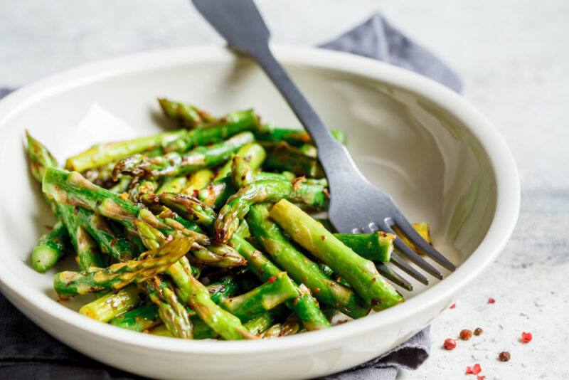 a white bowl with roasted green asparagus with silver fork resting on a grey table napkin