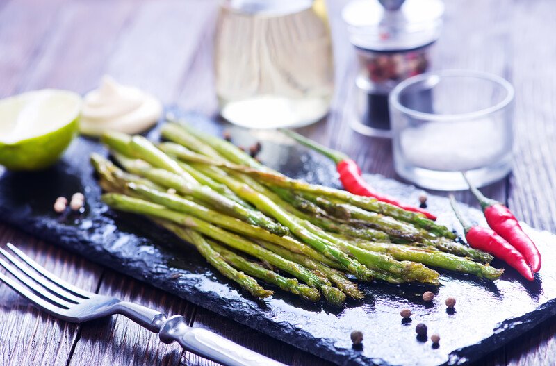 a closeup image of a rectangular black slab of rock with roasted asparagus on it with 3 red chilis, peppercorns, a mound of dipping sauce, and sliced lemons, beside it as a fork and a jar of oil and jar of peppercorns