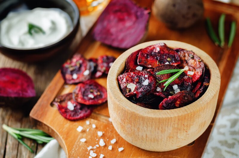 on a wooden chopping board is a wooden bowl with roasted beets with herbs, with loose roasted beets and rock salts beside it and a black bowl with white dipping sauce