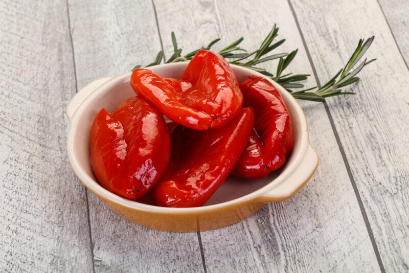 on an aged wooden surface is a gold and light brown bowl full of roasted bell peppers, with a sprig of fresh rosemary at the back