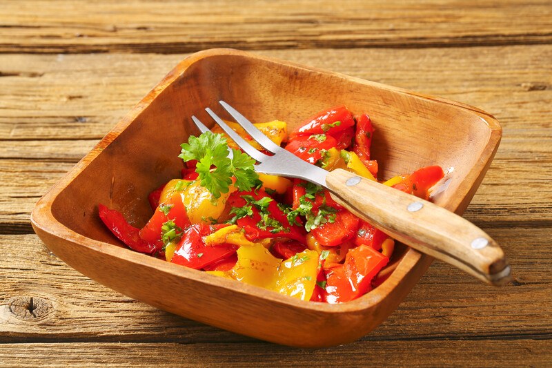 on a wooden surface is a rectangular bowl with roasted red and yellow bell peppers with herbs and a fork with wooden handle