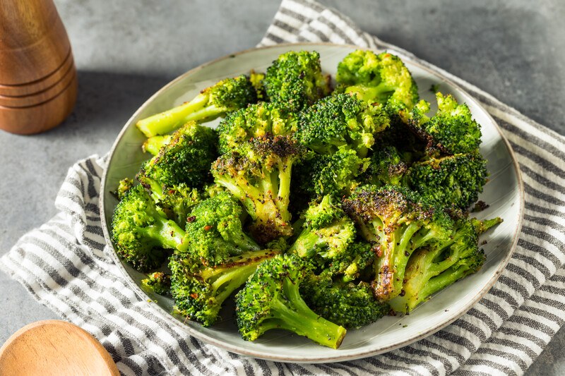 white plate full of roasted broccoli with a white and grey table napkin with partial image of wooden pepper mill and spoon