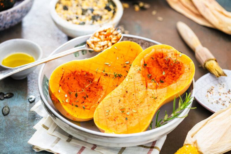 on a wooden surface is a white shallow bowl is a roasted halved butternut squash resting on a white napkin with brown stripes, around it are small dishes of olive oil and seeds, brush and loose seeds