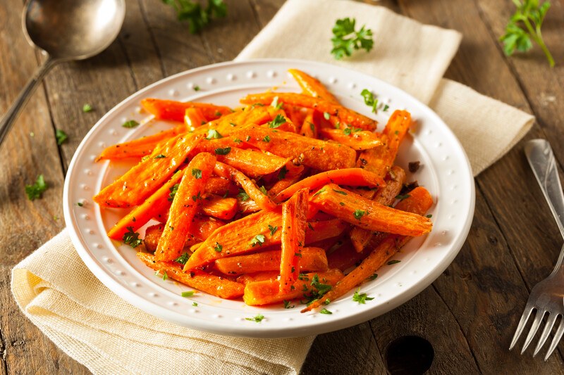 closeup image of a white ceramic dish with roasted carrots sticks with herbs on top, underneath is a white table napkin