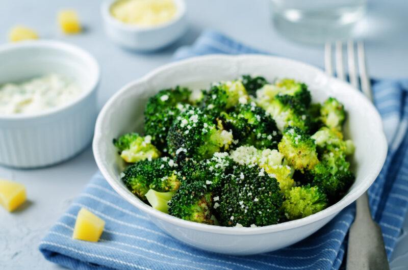 on a concrete surface is a white bowl of roasted cauliflower resting on top of a blue table napkin with white stripes, beside it is a silver fork and a couple of dishes with rock salt and yellow sauce, a glass of bottle, and loose cubed potatoes around it