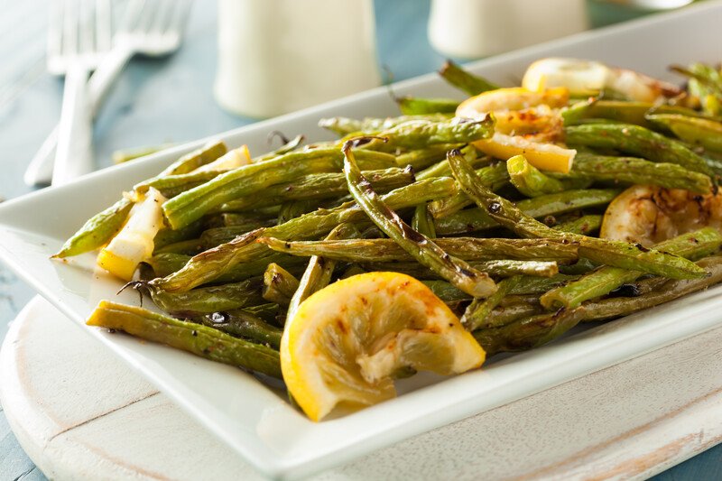 partial image of a rectangular white dish with roasted green beans with lemon slices, resting on top of a white round wooden board