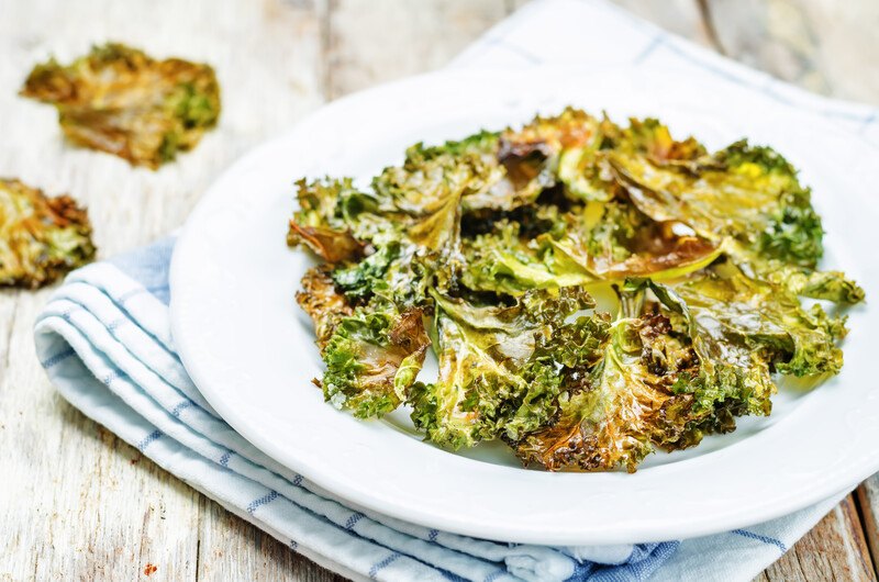 on an aged wooden surface is a white dish full of roasted kale on top of a striped white and dark green table napkin with a couple of loose roasted kale at the back